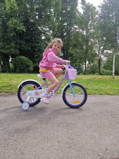 A young girl is riding a purple and white bicycle with training wheels on an asphalt path. She is wearing a pink hooded jacket and pink shorts. The bicycle has a purple basket on the handlebars filled with white and purple items. The background consists of green grass and trees. The image appears to have been taken on a bright, overcast day. The setting is a park or recreational area. There is no visible indication that this location is in the city of Salford, United Kingdom.