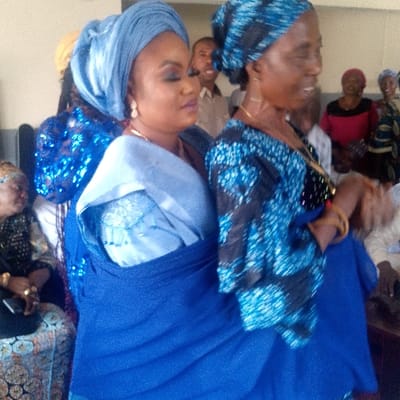 This image captures an indoor social gathering in Ozuri, Nigeria. The foreground is dominated by two women dressed in shades of blue. The woman on the left wears a light blue gele (headwrap), a lighter blue patterned top, and a royal blue wrapper, accessorized with gold drop earrings and a gold necklace. Her facial features include noticeable makeup, and she looks towards her right. The woman on the right wears a blue and black patterned headwrap and a matching top over a royal blue skirt. She has a gold chain necklace and gold bangles on her left wrist, and her gaze is directed towards her right.
In the blurred background, numerous other individuals are present, indicating a crowded event. A man with a smile is visible centrally in the mid-background. Other attendees include a person partially obscured on the far left wearing a dark, possibly sequined, blue top, and several women in various attire, such as a red top and a yellow headwrap, seated or standing. The environment consists of light-colored walls and indistinct furniture, suggesting a functional interior space.