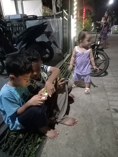 Two boys are sitting on a drain grate looking at a mobile phone. A baby stands nearby wearing purple pajamas and shoes. A motorbike is parked next to the grate. An adult woman in a floral dress and another baby can be seen in the background behind the parked bicycle. The location is Bungur, Indonesia. The background shows buildings, a gate, and lighting.