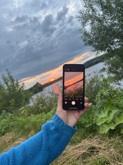 A hand wearing a blue knitted sweater holds a smartphone, capturing a sunset over a body of water. The phone's screen displays a camera interface with various shooting modes, including "Portret" (Portrait), "Foto" (Photo), and "Video" (Video). The image on the phone shows the sun setting on the horizon, casting orange and red hues across the sky and reflecting on the water. Trees with green foliage line the opposite shore, and dark clouds are visible in the upper left portion of the scene. The overall setting appears to be outdoors, near a river or lake, with lush greenery and dry grass in the foreground. The time of day is clearly sunset, indicated by the warm colors in the sky.