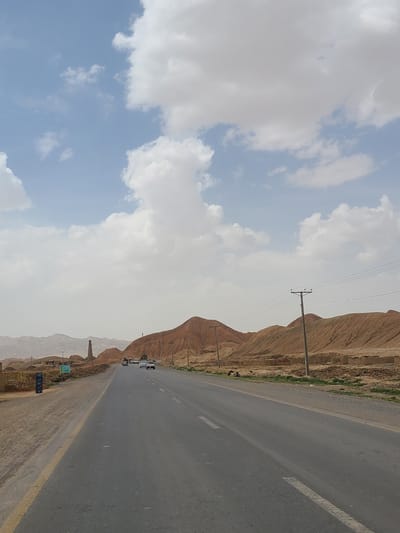 The image depicts a roadside view in an arid, mountainous region, likely located in Pakistan. A paved highway runs along the right side of the frame, displaying yellow and white lane markings. The highway leads toward distant mountains. The mountains appear barren, with hints of vegetation near the road. Power lines run along the mountains, supported by utility poles. Several cars are visible on the road. The left side of the frame showcases a sky with white and grey clouds. The exact location is Unknown, Pakistan.