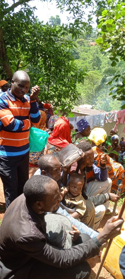 A group of people gather outdoors. A man in an orange and blue striped shirt stands while others sit on the ground. A child sits on someone's lap, and several people are wearing colorful head coverings. Laundry is hanging on a line in the background. The group appears to be in a rural setting, possibly a village, with lush green vegetation surrounding them.