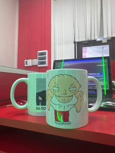 The image depicts two mugs on a red table. The mug in the foreground features a cartoon character with an angry expression, muscular arms, and is wearing a white tank top and red pants. The mug to the left is lighter in color and has a photo or graphic of a person's arm and hand resting on a dark surface. Green laser lines are visible crossing both mugs and a laptop screen in the background. The background shows a doorway with vertical blinds and a sign that reads "ВЫХОД" (EXIT in Russian). Electrical boxes are mounted on a red wall to the left. The scene is indoors, possibly an office or a room with a desk. The location of Yegorlykskaya, Russia is not evident from the visual information in the image.