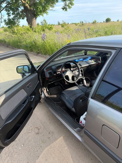 The image shows the interior of a gray car with the driver's door and passenger door open. The car is parked on a dirt road in a rural area, with a field of tall grass and purple wildflowers to the right and a large tree to the left. The interior of the car is visible, including the dashboard, steering wheel, and seats. There are two blue packs of Oreos on the dashboard and a black air freshener hanging from the rearview mirror. A white plastic bag with red lettering is on the floor of the passenger side. The car appears to be in good condition. The image was taken during the daytime, with clear blue skies and some clouds. The location is described as Povarovka, Russia.