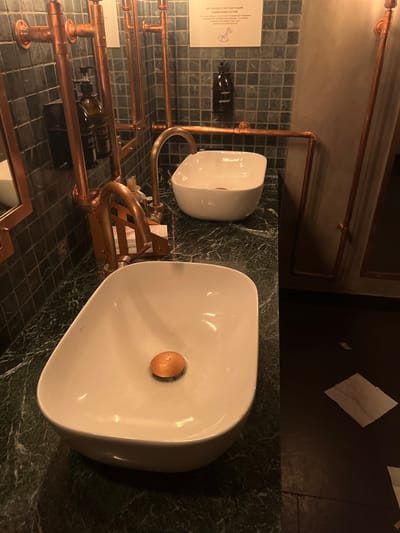 In a bathroom with dark tiled walls and a dark green marble countertop, two white ceramic sinks are visible. The sink in the foreground has a copper drain stopper. Above the sinks, copper pipes form a utilitarian faucet system. To the left, a brown glass bottle with a pump dispenser, labeled with Cyrillic text, is mounted on the wall. A small, white sign with Cyrillic text and a drawing of a giraffe is attached to the wall above the bottles. The floor is dark, and a few crumpled white paper items are scattered on it. The overall impression is one of a rustic or industrial-chic design. This scene is located in Vyshen'ky, Ukraine.