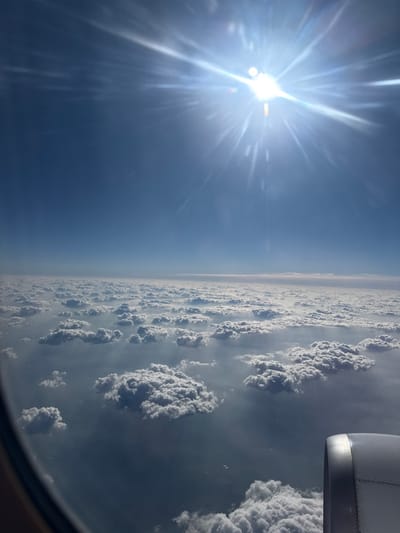 This image captures an aerial view from an aircraft window, depicting a high-altitude scene during a flight associated with the city of Villeparisis, France. The upper half of the frame shows a clear, bright blue sky. The sun, located in the upper right quadrant, is intensely bright, characterized by significant lens flare and radial light rays. Below the clear sky, a dense and expansive layer of cumulus clouds stretches horizontally, forming an uneven landscape of white and grey masses. A faint, hazy horizon line separates the cloud layer from the upper sky. The lower left portion of the image reveals the curved, dark frame of the aircraft's window, while the lower right displays a metallic, silver-grey section of an aircraft engine nacelle. No discernible human figures, specific ground features, or overt actions are depicted. The scene is static, documenting atmospheric conditions and parts of the aircraft from which it was observed.