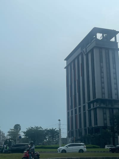 In Pakulonan Barat, Indonesia, a tall, modern building with a dark facade and vertical orange accents dominates the skyline under a hazy, overcast sky. The building features multiple stories with rows of windows and a distinctive, angled roof structure. In the foreground, a road runs parallel to a grassy median. Several motorcycles are on the road, including one red scooter with a rider wearing a helmet and a backpack. A white minivan is also visible on the road. Behind the median, a line of trees forms a backdrop to the scene, with the sky completely obscured by a light fog or haze. The overall atmosphere is muted and somewhat diffused due to the atmospheric conditions.