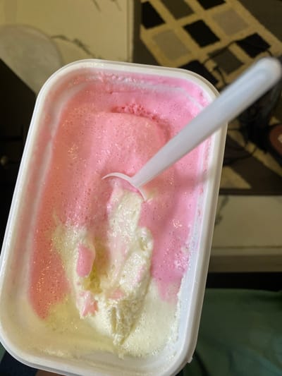 The image shows a close-up, overhead view of a white rectangular container filled with ice cream. The ice cream is a swirl of pink and white, with a white plastic spoon embedded in it, suggesting it has been eaten. The container is slightly tilted. The background is blurred, showing a patterned rug on the floor and a dark green cloth. There is no indication of the city of Dam Camp, Nigeria, in the image.