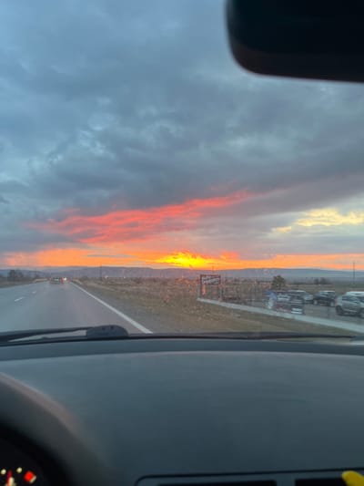 The image presents a roadside scene at either sunrise or sunset, viewed from the interior of a vehicle. The sky is dominated by a band of intense orange and yellow light along the horizon, indicative of the setting or rising sun, transitioning into dense, dark gray clouds covering the upper sky. A wet, two-lane asphalt road with a broken white center line and a solid white shoulder line extends into the distance. Two vehicles are visible ahead in the same direction, their taillights illuminated. To the right of the road, a commercial sign with Cyrillic script reads "AUTO Daskalov," positioned adjacent to a lot containing several parked vehicles, including cars and SUVs. The surrounding environment consists of sparse, dry vegetation and fields, with distant hills or mountains visible along the horizon. The foreground shows the dark dashboard and a segment of the steering wheel/instrument cluster of the originating vehicle. The scene is located in Krainitsi, Bulgaria.