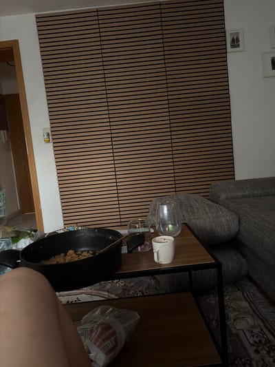 This media file shows an interior scene with a wooden slatted wall and a corner of a couch. On a wooden table, there are two glasses, one containing a liquid and the other a white rolled-up item, possibly a napkin or paper towel. To the right, a black frying pan with food inside is visible, next to a bag with text on it. A person's leg is also visible in the lower right corner. The lighting suggests it might be evening or a dimly lit room. The scene appears to be in a domestic setting, possibly a living room or kitchen area, given the furniture and household items. The presence of the frying pan suggests a meal may have been recently prepared or is being consumed. No specific weather conditions or emotions are discernible.