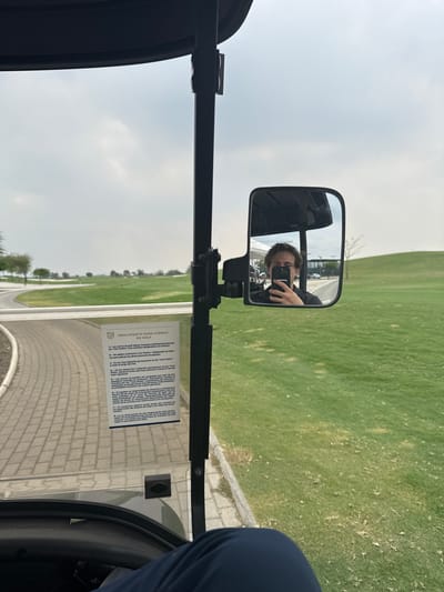 A person is reflected in the side mirror of a golf cart, holding a phone to take a picture. The cart is driving on a paved path alongside a golf course, with green grass visible. A rules sheet in Spanish is attached to the cart's side. The sky is overcast.