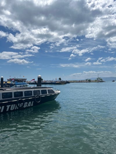 The image depicts a harbor scene in the city of Tidore Islands, Indonesia, under a bright, partly cloudy sky. In the foreground, a black and white passenger boat, identified by the text "RI TUNGGAL" on its side, is docked in the calm, blue-green water. The boat has multiple windows and two black spare tires hanging from its hull. Behind this boat, several other passenger boats are lined up, many adorned with small flags. Further in the background, a small, rocky island with sparse vegetation and a lamppost stands in the sea, with a mountainous coastline visible in the distance. The water appears clear and reflects the sky and surrounding boats. The overall scene is one of a bustling port on a sunny day.