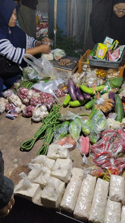 The image depicts a market stall, likely in Sukodono, Indonesia, with a variety of fresh produce and packaged goods. Two individuals are partially visible, one woman wearing a blue hijab and a striped shirt is attending to items on a scale. She appears to be interacting with plastic bags of produce. The other person, seen from behind, wears a black hijab and glasses, and is looking down. The stall is laden with vegetables such as garlic, shallots, long beans, chilies, eggplants, and cucumbers. There are also several blocks of what appear to be tofu or tempeh wrapped in plastic, and small packets of spices or seasonings. The lighting suggests it might be evening or a dimly lit indoor market. The overall atmosphere is that of a bustling local marketplace. The scene is rich with the colors and textures of fresh food.