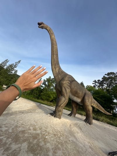 An outdoor, eye-level, wide shot shows a large, brown, dinosaur statue, likely a Brachiosaurus, with a long neck and tail, standing on a light-colored, sandy surface. The dinosaur is positioned in the center-right of the frame, with its head reaching towards the top of the image. In the foreground on the left, a person's left arm and hand are visible, with the hand outstretched towards the dinosaur, fingers splayed. The person is wearing a green bracelet on their wrist. The background consists of a bright blue sky with some wispy white clouds, and lush green trees and foliage. The location is identified as Baan Lipa Noi, Thailand.
