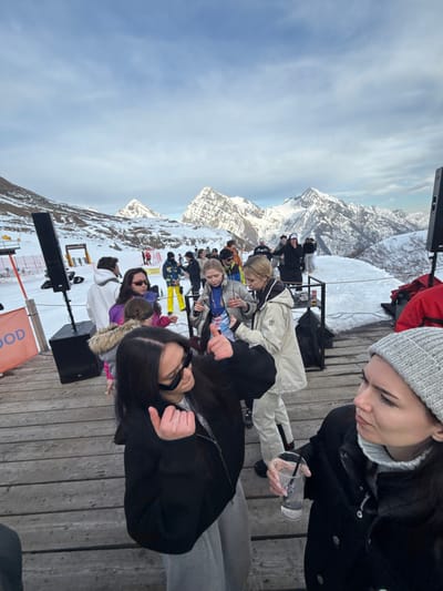 The image depicts an outdoor social gathering on a wooden deck in a snow-covered alpine setting, identified as Alagna Valsesia, Italy. The bright daytime scene is dominated by majestic snow-capped mountains under a partly cloudy sky.
In the foreground, two young women are prominent. On the right, a woman in a grey beanie and black winter jacket holds a clear plastic cup with ice, looking down introspectively. To her left, another woman with long dark hair, wearing sunglasses and a black jacket, is partially visible, making a hand gesture with both thumbs pointing downwards. Behind them, several people, mostly young adults dressed in ski wear and various winter clothing, are socializing, some seated and others standing.
A large black speaker on a stand is positioned on the wooden deck in the mid-ground, with another speaker partially visible further back. The deck itself is constructed from wooden planks. The setting is a high-altitude terrace or viewing platform amidst a vast, rugged mountain range, typical of a ski resort.
The activity appears to be an apres-ski event or an outdoor party/concert, characterized by the presence of music equipment and people enjoying drinks and conversation in the chilly environment. The weather is cold and clear, with abundant snow covering the ground and peaks. The time of day is daylight. The woman in the beanie shows a pensive expression, while the other woman appears more animated. The location cues strongly suggest a winter sports destination.
The word "WOOD" is partially visible in white lettering on an orange sign or box near one of the speakers on the deck.