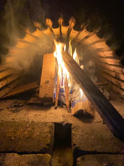 A fire burns within a circular structure made of curved, clay-like bricks. The fire is fueled by wooden planks and cardboard. Smoke emanates from the flames. The fire pit is positioned on a surface of concrete bricks. The location is unknown; the city of Konstantinovo, Bulgaria is not visible in the image. There are no people present in the scene.