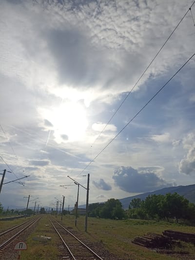 The image depicts a railway scene in Bliznets, Bulgaria. Two sets of parallel train tracks stretch into the distance, flanked by grassy verges and utility poles with overhead power lines. To the right of the tracks is an area of scrubland with several stacks of logs. Beyond the scrubland is a line of trees and a distant mountain range. In the lower-left corner, a red and white circular sign is visible. The sky is filled with clouds, and the sun is obscured, creating a diffused light. There are no people visible.