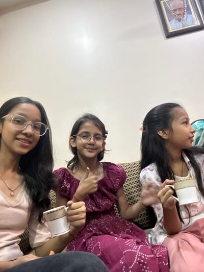 Three girls are casually seated on a patterned couch. One girl wears a pink dress, eyeglasses and a necklace, another wears a purple patterned dress and eyeglasses, and the third wears a white patterned top. All are holding teacups. A framed picture hangs on the wall. The image is rotated counterclockwise.