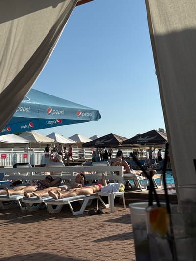 The image depicts an outdoor pool area with several people relaxing on lounge chairs. Two large blue Pepsi umbrellas are prominently displayed in the background. Some individuals are sunbathing, while others are standing or walking. A swimming pool is visible to the right, with blue water. The ground is paved with brick-like tiles. The scene appears to be in a resort or public pool facility. The location is identified as Luka-Meleshkivska, Ukraine.