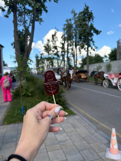 A close-up, low-angle shot shows a hand holding a dark red, intricately shaped lollipop resembling a Matryoshka doll. The lollipop is on a thin wooden stick. The hand has long, manicured fingernails painted white and is wearing a simple silver ring on the index finger. The background is blurred but suggests an outdoor urban environment with trees, a paved walkway, and a road. In the middle ground, several horse-drawn carriages are visible, some with people on them, implying a tourist attraction or event. A person in bright pink clothing with a backpack is in the left background. Further back, buildings and more trees line a street. The sky is blue with scattered white clouds. The scene appears to be in the city of Suzdal, Russia, known for its historical architecture and horse-drawn carriage rides.