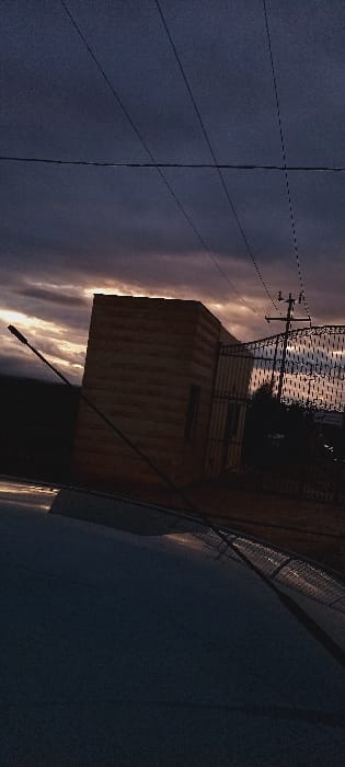 A dimly lit outdoor scene showcases a tan, rectangular building, possibly a container or small structure, set against a twilight sky streaked with orange hues. A metal fence surrounds the rooftop. Power lines stretch across the top left corner. A street light pole extends upwards from the bottom, hinting at a street-side location. The bottom of the image has a darker, bluish tone, suggesting pavement or ground cover.