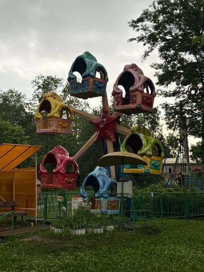 The image shows a small, faded amusement park ride, possibly a carousel or a miniature Ferris wheel, with several gondolas designed in the shape of octopus heads. The ride is situated in an outdoor, grassy area, with trees in the background and a cloudy sky overhead. To the left, there is an orange awning and a wooden bench. Fencing encloses the ride. In the background, a building and other amusement park structures are visible. The overall impression is one of a slightly neglected, old-fashioned amusement park. The ride appears to be unoccupied. The image does not contain any specific details to confirm its location in Chusovoy, Russia, as it is a generic scene of an amusement park.