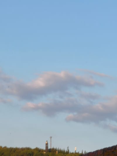 The image shows a wide shot of the sky over Passignano sul Trasimeno, Italy. The sky is clear and blue with some scattered clouds. In the distance, a treeline can be seen, with a church tower peeking out from behind the trees. The sun appears to be setting, casting a warm glow over the scene. The image is taken from a high vantage point, likely from a hill or building overlooking the town.