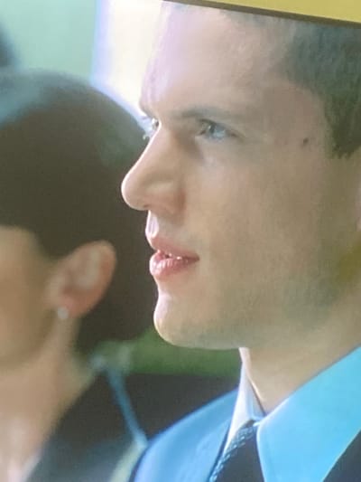 A close-up, rotated image captures a section of a person's face, specifically from the eye down to the chin, and part of their neck and shoulder, alongside a blurred hand in the foreground. The person, who appears to have light to medium skin tone, is seen in profile with their head tilted. A light-colored eye (possibly blue or grey) is visible, and there's a small dark spot on the cheek. Short, dark hair is visible at the top left. The lips are slightly parted, revealing some teeth. Below the chin, the outline of a jawline with what appears to be light stubble is discernible.
On the right side of the frame, part of a light blue collar and a dark, possibly patterned, tie or strap are visible, suggesting formal or semi-formal attire. In the lower right corner, a blurry hand is present, with a ring visible on one of the fingers, possibly the pinky or ring finger. The background is indistinct and out of focus, showing blurry patches of green and dark tones. The lighting is soft, and there are no clear indications of the time of day or specific weather conditions. No visible text is present. The overall scene gives the impression of a moment captured during speech or deep thought, likely from a video or television screen due to the image quality. This scene is contextualized as being in Gosa, Nigeria.