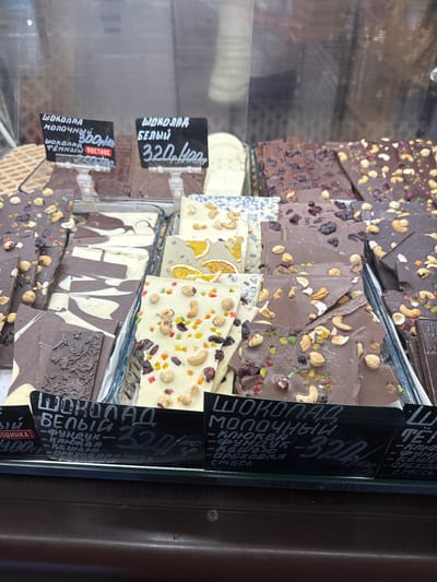 The image shows a close-up of a display case filled with various types of artisan chocolate bars. The chocolates are arranged in stacks and on trays, with visible ingredients like nuts, dried fruits, and candied citrus peel embedded in them. Some bars have intricate designs etched into their surface.
Several black price tags are attached to the display, with handwritten text in Cyrillic. One tag reads "ШОКОЛАД БЕЛЫЙ 320р." (White Chocolate 320 rub.). Another tag indicates "ШОКОЛАД ТЁМНЫЙ ПОСТНОЕ 300р." (Dark Chocolate Vegan 300 rub.). A larger tag further down lists "ШОКОЛАД МОЛОЧНЫЙ" (Milk Chocolate) with ingredients like "ФУНДУК" (hazelnut), "КЛЮКВА" (cranberry), "КЕШЬЮ" (cashew), and "ОРЕХОВАЯ СМЕСЬ" (nut mix), also priced at 320.
The overall scene suggests a confectionery shop or market stall. The lighting appears to be indoor, with reflections visible on the glass display case. The chocolates vary in color, from deep dark brown to light cream. The detailed textures and ingredients suggest high-quality, handcrafted products. The context provided indicates the location is Svkh Maslovskiy 1 Otdeleniye, Russia. No people are visible in the image.
