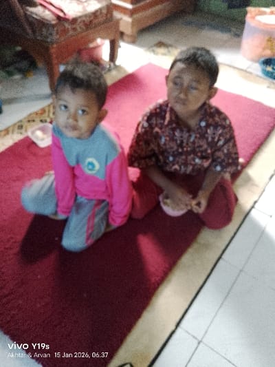 Two young children sit on a maroon rug indoors. The child on the left wears a pink and grey outfit, gazing directly at the camera. The child on the right, in a patterned shirt and reddish pants, holds a small dish. Both are seated on their knees. The rug is placed atop light-colored tiles. Furniture is visible in the blurred background, including a cushioned chair or ottoman and a cabinet. A pink bucket sits in the far background. The bottom of the frame contains a watermark and date.