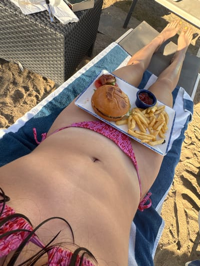 A woman in a pink bikini lies on a beach chair with a striped towel, displaying a plate of food. The plate holds a burger, french fries, and a small bowl of ketchup. The scene is outdoors, likely at the beach. The chair and the woman are in the sunlight, with sand visible in the background.