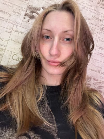 This is a close-up, selfie-style photograph of a young woman with light-colored, long, wavy hair, featuring streaks of blonde and reddish-brown. She is looking towards the camera with a neutral expression. Her eyes are blue and she has a small nose ring. The background is a textured wallpaper with a pattern of coffee-related text and imagery, including words like "Mundo Noro," "Mocha," "Arabica," and "Sumatra." The lighting suggests an indoor setting, likely during the daytime. The woman is wearing a dark t-shirt with a faint graphic design. The overall impression is casual and personal.