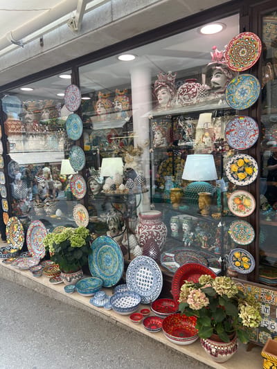 This is a street-level view of a shop window in Taormina, Italy, displaying a vibrant collection of ceramic art. The window is filled with hand-painted ceramics, including large decorative plates featuring intricate floral and geometric patterns, as well as depictions of fruits and human faces. Several ceramic heads, adorned with ornate crowns and detailed facial features, are prominently displayed. Among the ceramics, there are also table lamps with light blue shades and decorative vases in various sizes and designs, some with intricate burgundy patterns and others with face motifs. A lush green plant with light green hydrangea blooms is placed in the foreground, adding a natural element to the display. The ceramics are arranged on shelves and on the ledge outside the window. The reflection on the glass shows a street and possibly other buildings, suggesting an outdoor setting.
