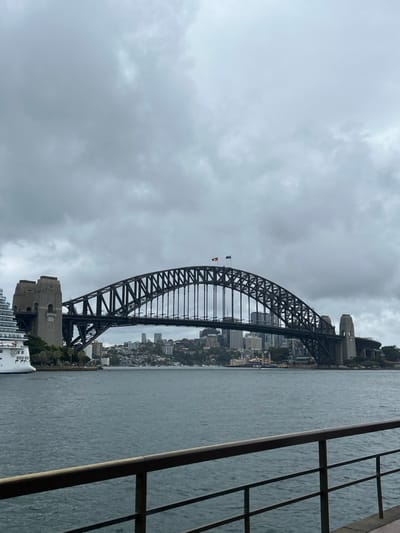 Sydney Harbour Bridge dominates the view under an overcast sky. The bridge's dark silhouette contrasts with the gray sky and water. Skyscrapers line the harbor behind the bridge. A structure with metal railings is partially visible on the right. A building complex can be seen on the lower left.