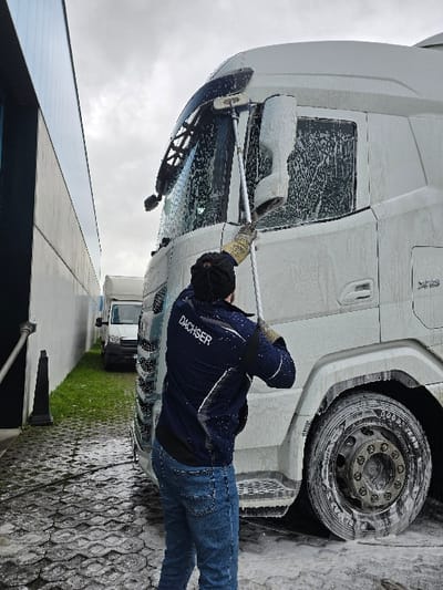 In the city of Ostend, Belgium, an individual is actively washing a large, white DAF XF semi-truck, which is heavily coated in white soap foam. The person is observed from behind, wearing a dark blue jacket with "DACHSER" emblazoned in white across the back, blue jeans, a dark beanie, and work gloves. They are holding a long, silver-handled washing brush, scrubbing the front windshield and upper cabin of the truck. The scene unfolds outdoors on a dark, wet, wave-patterned paved surface that shows traces of foam. A tall, dark corrugated metal building wall runs along the left side of the frame. In the background, a narrow alleyway between this wall and another lighter-colored wall reveals a strip of green grass and two parked white vans. The sky above is overcast and grey. A black, truncated traffic cone is positioned on the ground near the left wall.