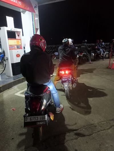 The image depicts a nighttime scene at a gas station (SPBU) in Cimaung, Indonesia, where several motorcyclists are queued up to refuel. The dark sky confirms the time of day, with the area illuminated by the gas station's artificial lights.

In the foreground, a person is seated on a black scooter, viewed from behind. They are wearing a red full-face helmet with white and yellow designs, a black jacket, blue jeans, and white sneakers with red accents. The scooter's license plate, "D 6153 VBF," is clearly visible, indicating an expiration date of "07-28" (July 2028). Directly in front, another individual on a scooter also waits, wearing a dark hoodie, yellow shorts, and a patterned helmet. Their scooter's illuminated red taillights are visible, as is its license plate, "D 5658 ZAK," with an expiration date of "05-27" (May 2027). Long shadows of the motorcycles and people are cast across the concrete ground.

To the left, a white and red fuel dispenser stands prominently, displaying "PERTAMAX 92," the "PERTAMINA" logo, and "PASTI PAS" text, identifying the fuel brand and type. In the mid-ground and background, more motorcyclists are seen waiting in line. A white metal fence runs horizontally in the background, and a red and white striped traffic cone is partially visible on the far right. The scene portrays a routine evening activity at a busy fueling station.
