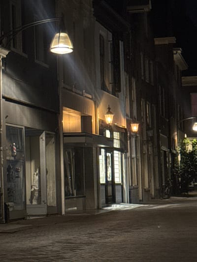 The image depicts a nighttime street scene in Deventer, Netherlands. The street is paved with cobblestones, and the surrounding buildings are lit by ornate streetlights and interior lights. Several shops are visible, with large glass windows displaying their interiors. One shop appears to be a barber shop, identifiable by a barber pole. Another shop window reveals a mannequin or statue dressed in clothing. Trees are present on the right side of the street, and a bicycle is parked near one of the buildings. The atmosphere is quiet and dimly lit, suggesting a late hour.