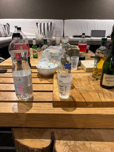 A wooden coffee table displays an array of beverages: a champagne bottle, a Coca-Cola bottle, an Absolut vodka bottle, two water bottles labeled "OLA AGUA," and several Stella Artois beer bottles. A bowl filled with ice and plastic cups is also visible. The table is positioned near a white sofa, with a television in the background. A striped pillow rests on the sofa. The scene suggests a social gathering or a casual setting.