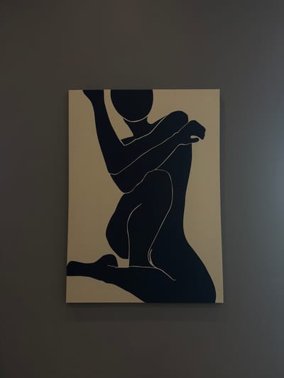 A framed canvas print hangs on a dark gray wall. The artwork depicts two figures in a modern, minimalist style. The figures are rendered in solid black against a tan or beige background. Their bodies are intertwined. The canvas is rectangular and appears to be hung evenly on the wall. The wall itself has a slightly textured surface. The location is Palm Desert, United States of America.
