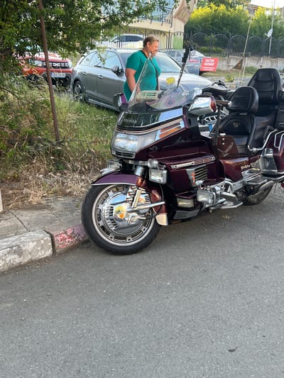 The image shows a close-up of a maroon Honda Gold Wing motorcycle parked on an asphalt road. The motorcycle is adorned with chrome accents, including its wheels, engine components, and handlebars. The front wheel features a prominent chrome rim with intricate detailing, and a circular chrome cover in the center. The motorcycle's plush leather seats are visible, as well as a windshield with a decorative decal.
In the background, a woman in a teal top is partially visible, leaning over the motorcycle. Behind her, two cars are parked: a grey Audi SUV and a bright orange Dacia Duster with a Bulgarian license plate reading "CB 136 XK." The scene appears to be in a semi-urban or residential area, with overgrown vegetation and a concrete curb running along the edge of the road. A red sign with white lettering, which reads "ЧАСТЕН ПАРКИНГ" (Private Parking), is also visible in the background. This suggests the location might be near a commercial establishment or private property. The overall lighting indicates it is daytime, with soft sunlight casting gentle shadows.