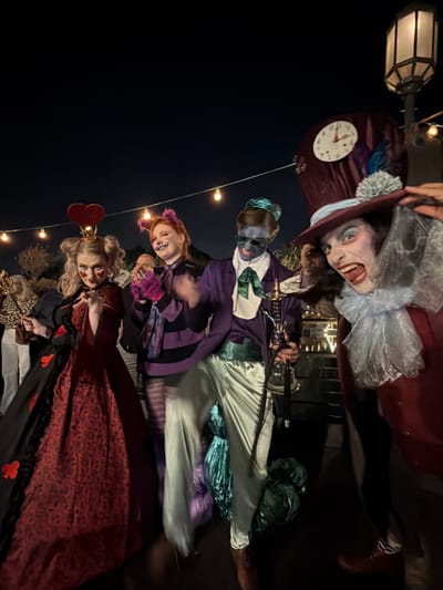 This image captures four individuals dressed in elaborate fantasy-themed costumes, likely at an outdoor evening event in Nir Eliyahu, Israel. The scene is set at night, with the subjects illuminated by artificial string lights and a streetlamp visible on the left.
From left to right, the costumes appear to be inspired by characters from "Alice in Wonderland" or similar fantasy settings:
1. **Mad Hatter/Joker-like figure:** This person wears a tall, maroon top hat decorated with a clock face, a fluffy white pom-pom, and a blue feather. Their face is painted white with dark eye makeup and a wide, theatrical grin, complemented by a white ruffled collar and a maroon coat.
2. **Blue-faced character:** Next to them is a figure with blue face paint, dressed in a purple top with white accents and light-colored trousers. They appear to be holding a prop weapon, possibly a sword or staff.
3. **Cheshire Cat-like figure:** This individual has orange-red hair with cat ears, a purple and black striped outfit with furry pink details on the sleeves, and a wide, painted smile.
4. **Queen of Hearts:** On the far right, a person is costumed as the Queen of Hearts, featuring blonde hair, a golden crown with a red heart, a red and black patterned gown, and heart-shaped makeup on their cheeks. They are holding what appears to be a scepter.
The atmosphere is festive and theatrical, suggesting a themed party, Halloween celebration, or performance. The dark background highlights the brightly lit costumes and the expressive poses of the participants. The image is taken outdoors, indicated by the visible streetlamp and string lights. No readable text is visible other than the clock face on the hat.