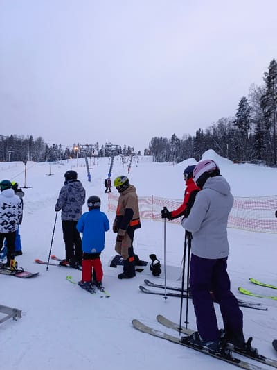 This image captures a group of people at the base of a ski slope, likely at a ski resort, on an overcast winter day. The sky is a uniform, light grey, suggesting either early morning, late afternoon, or consistently cloudy weather, with no direct sunlight visible.
The ground is entirely covered in white snow, indicating a cold, wintry environment. In the background, snow-covered evergreen trees line the hills, characteristic of a mountainous or hilly ski area.
Several ski lift pylons, appearing to be for T-bar or button lifts, ascend the slope in the mid-ground. Lights are visible on some of these pylons, suggesting the area might be lit for night skiing or for improved visibility during low-light conditions. Orange safety netting runs along the sides of the slope, separating areas or indicating boundaries. Other skiers and snowboarders can be seen further up the slope near the lift.
In the foreground, a group of approximately six individuals are gathered. They are all dressed in full winter sports gear, including helmets, ski jackets, and ski pants, indicating they are prepared for skiing or snowboarding. Most are wearing skis, while at least two individuals are on snowboards.
From left to right:
* A person in a white and black patterned jacket and black helmet stands on a snowboard, facing away from the camera.
* Next to them, a person in a grey and white patterned jacket and black helmet stands on skis, holding ski poles, also facing away.
* A younger individual, likely a child or teenager, in a bright blue jacket, red pants, and black helmet, is on skis and facing away.
* An adult in a tan and black ski/snowboard suit with orange accents and a green helmet stands on a snowboard, looking to the left.
* Further to the right, two people are standing on skis, holding poles. One wears a red jacket and dark helmet, while the other, slightly in front, wears a light grey jacket, dark purple pants, and a pink helmet. Both are facing to the right.
* A pair of skis and ski poles are partially visible on the far right, suggesting another person just out of frame.
The group appears to be waiting in line for the ski lift or perhaps regrouping at the bottom of a run. The atmosphere seems calm, typical of people enjoying a winter sports outing. No discernible text is visible anywhere in the image.