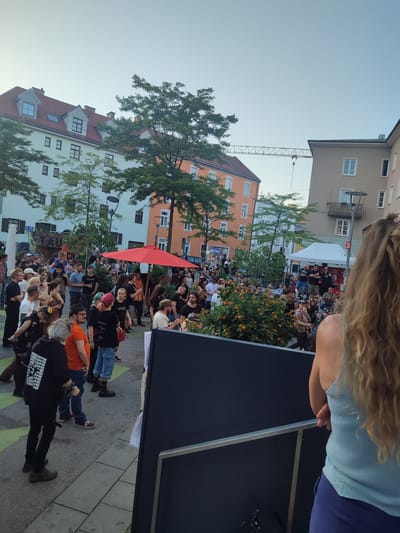In Rosenheim, Germany, a crowd of people gathers outdoors in a public square, likely for an event or performance. Buildings with multi-story facades line the background, one prominently featuring a red tiled roof and dormer windows. Trees with green foliage are scattered throughout the scene, and a red umbrella stands out among the attendees. A portable tent structure is visible in the distance, suggesting vendor or information booths. A construction crane is also present in the skyline, indicating ongoing development. The foreground shows a dark, rectangular structure, possibly a stage or barrier, with a person standing near it, their back to the camera, wearing a light blue tank top and dark pants. The ground is paved with grey and green tiles.
