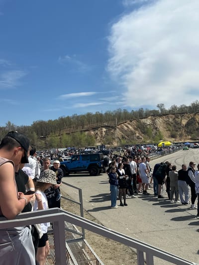 A crowd of people is gathered along a road or track, likely an event or race of some kind. A blue off-road vehicle is visible among the parked cars. There are trees on the other side of the road with a steep bank visible. The environment appears to be outdoors, likely a location in the Sadovoe, Russia area. A metal stairway-type structure with people sitting is to the right of the scene. The sky is blue with fluffy clouds.