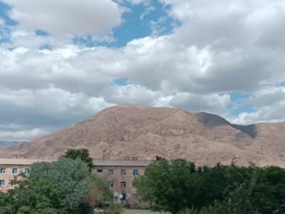 The image shows a vast, undulating brown mountain range under a sky filled with fluffy white clouds and patches of blue. In the foreground, a cluster of trees partially obscures two light-colored apartment buildings with multiple windows and satellite dishes. The buildings appear to be situated in a residential area, likely within the city of Balkanabat, Turkmenistan, given the characteristic arid mountain landscape and functional, utilitarian architecture common to the region. The scene is quiet and static, depicting a daytime view with natural lighting. There are no people or animals visible in the image.