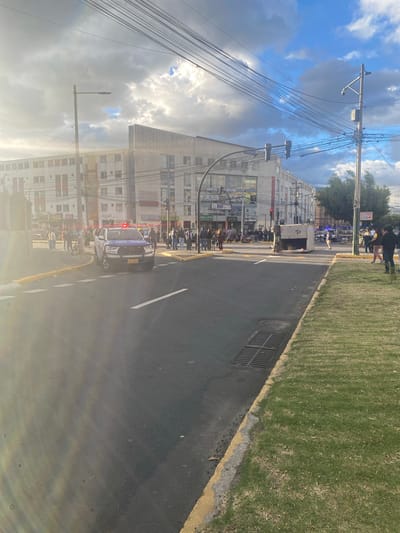 The image shows a street scene in Quito, Ecuador, likely during the daytime. A police vehicle with flashing lights is parked on the right side of the road, facing away from the camera. Several people are gathered around the vehicle and on the sidewalk.
The setting appears to be urban, with multi-story buildings lining the street. The buildings have many windows and are adorned with signs, some of which are partially visible. A large number of power lines crisscross the sky, creating a complex network above the street. The road itself is paved and marked with white lines. To the right of the road, there is a strip of green grass, bordered by a curb. A storm drain grate is visible on the pavement near the grass.
The sky is partly cloudy with patches of blue sky visible. The lighting suggests it is daytime, possibly late afternoon due to the angle of the sun and the bright, yet diffused, light. The overall mood is unclear, but the presence of a police vehicle and gathered individuals might indicate an incident or a routine patrol.
Some visible text includes "POLICIA" on the police vehicle and license plate that reads "TEZ-1571." Other signs on the buildings are too indistinct to read clearly. The image is oriented vertically, with the sky appearing at the top and the street running downwards.