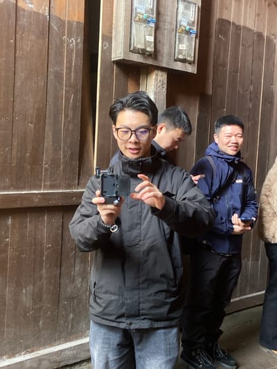 Three Asian men are indoors on a wood plank floor. One man is holding a camera and wearing glasses, a black jacket, and gray jeans. Another man is partially visible behind him wearing a dark jacket, and the third man is in the background wearing a blue jacket and black pants with boots. They are posing together for the photo. Wood planks line the walls.