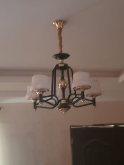 A chandelier hangs from a ceiling in a room. The chandelier has a central gold-colored stem wrapped in a brown material, from which black metal arms extend. Each arm supports a white lampshade with gold trim. The lampshades appear to be in the shape of upside-down cups. The ceiling is a neutral color. In the background, a dark, indistinct object, possibly a door or furniture, is visible. The image is blurry and lacks sharp detail. There are no people or actions depicted.