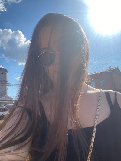 The image shows a young woman with long, brown hair, wearing sunglasses and a black top with a gold chain strap. She is taking a selfie outdoors on a sunny day. The background features a blue sky with scattered white clouds, a tall brick building with windows, and some electrical wires overhead. To the left, there is a distinctive cylindrical tower with circular windows, which is a known landmark in Poltava, Ukraine. The overall scene suggests a casual outdoor setting, possibly near a train station or industrial area, given the visible infrastructure. The sun is bright and high in the sky, casting a lens flare across the image.