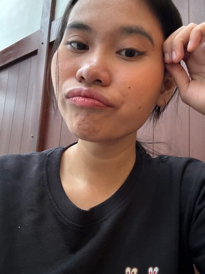 The image shows a close-up, selfie-style shot of a young woman with dark hair and brown eyes. She is wearing a black t-shirt and has her right hand raised, with her fingers touching her temple. Her lips are pursed, and she appears to be making a pouty expression. The background consists of a wooden wall with vertical slats. There is a faint reflection on her forehead, suggesting indoor lighting. The overall impression is of a casual, personal moment captured by the subject herself. The scene is likely indoors, possibly in a home or cafe, given the decor and lighting. There are no obvious indicators of a specific location within South Jakarta.